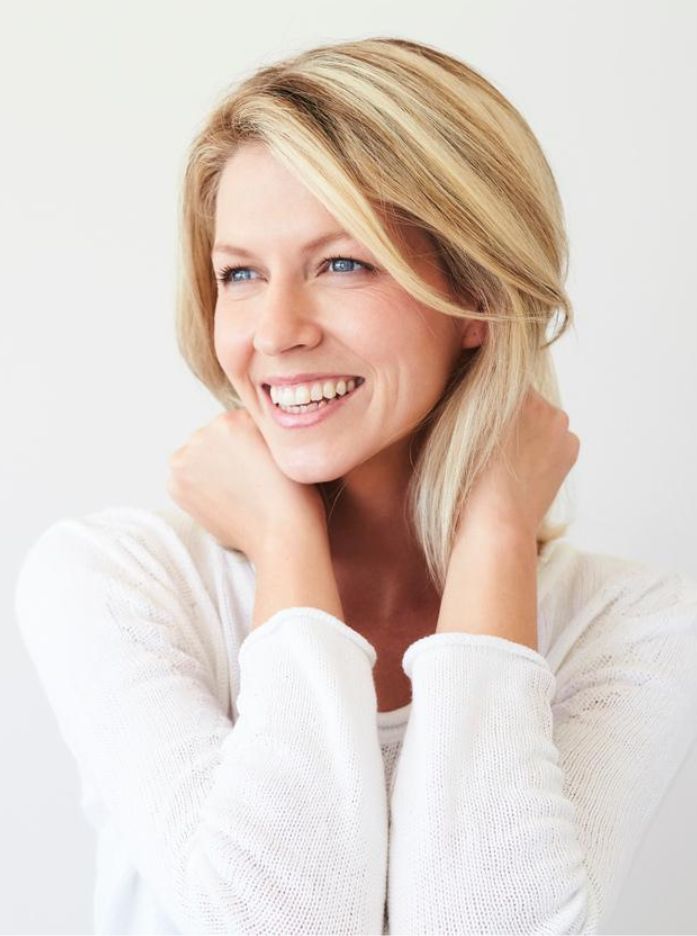 Smiling blonde woman in a white sweater looking right with hands near her neck