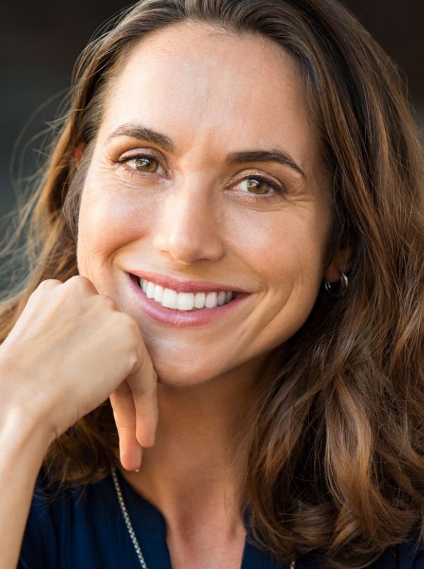 Smiling woman with shoulder-length brown hair resting chin on hand, outdoor headshot