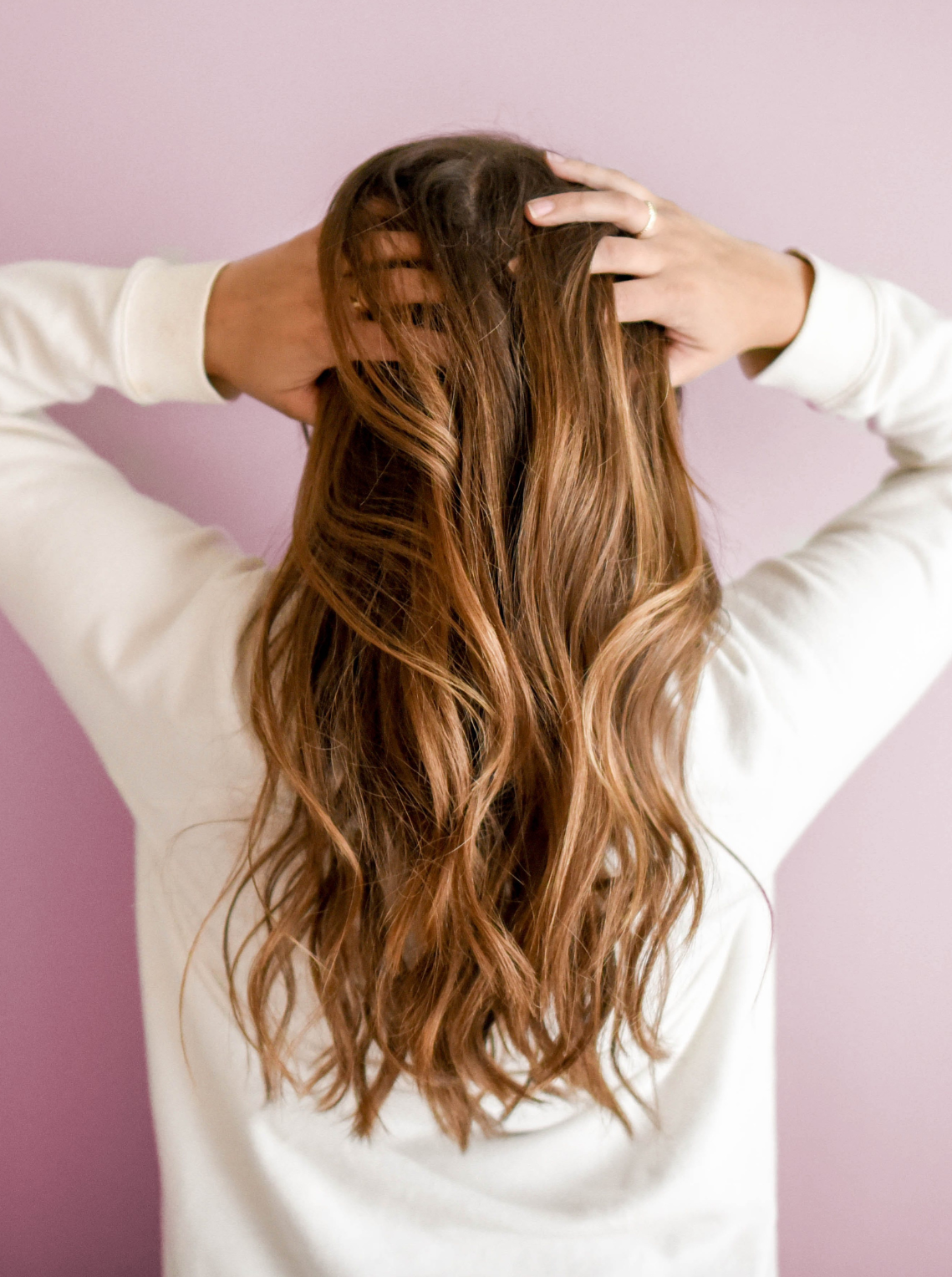 Woman shown from behind with long wavy brown hair, hands lifting hair against a pink wall