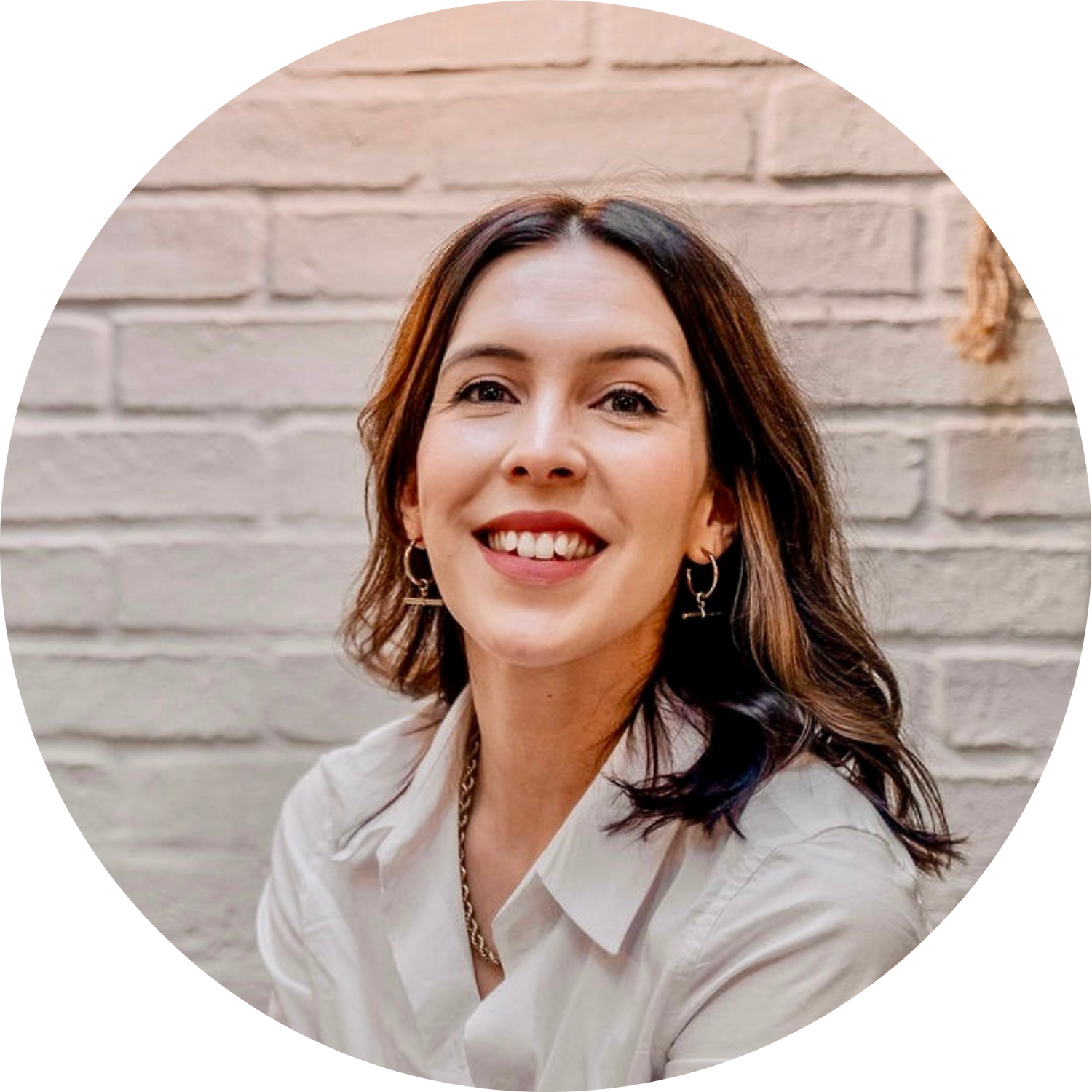Smiling woman with shoulder-length brown hair, wearing a white shirt and hoop earrings, against a light brick wall.